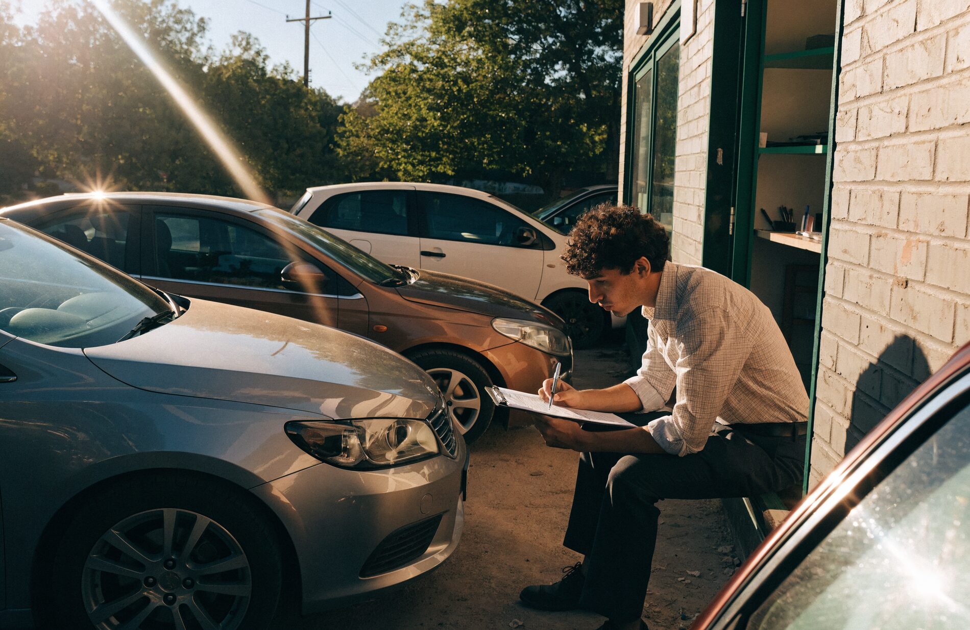 A dealer working through licensing paperwork on a clipboard beside a row of used cars on a small lot